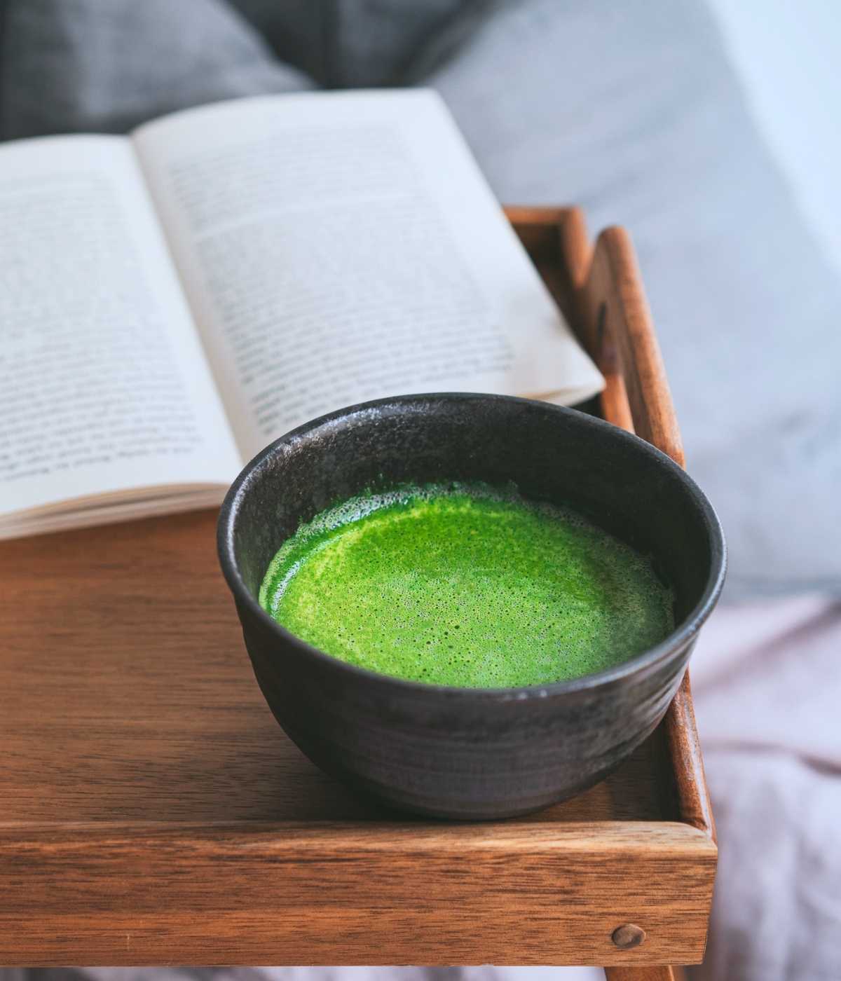 Matcha-Schale mit reinem Matcha-Pulver neben einem aufgeschlagenen Buch auf einem Nachttisch im Schlafzimmer.
