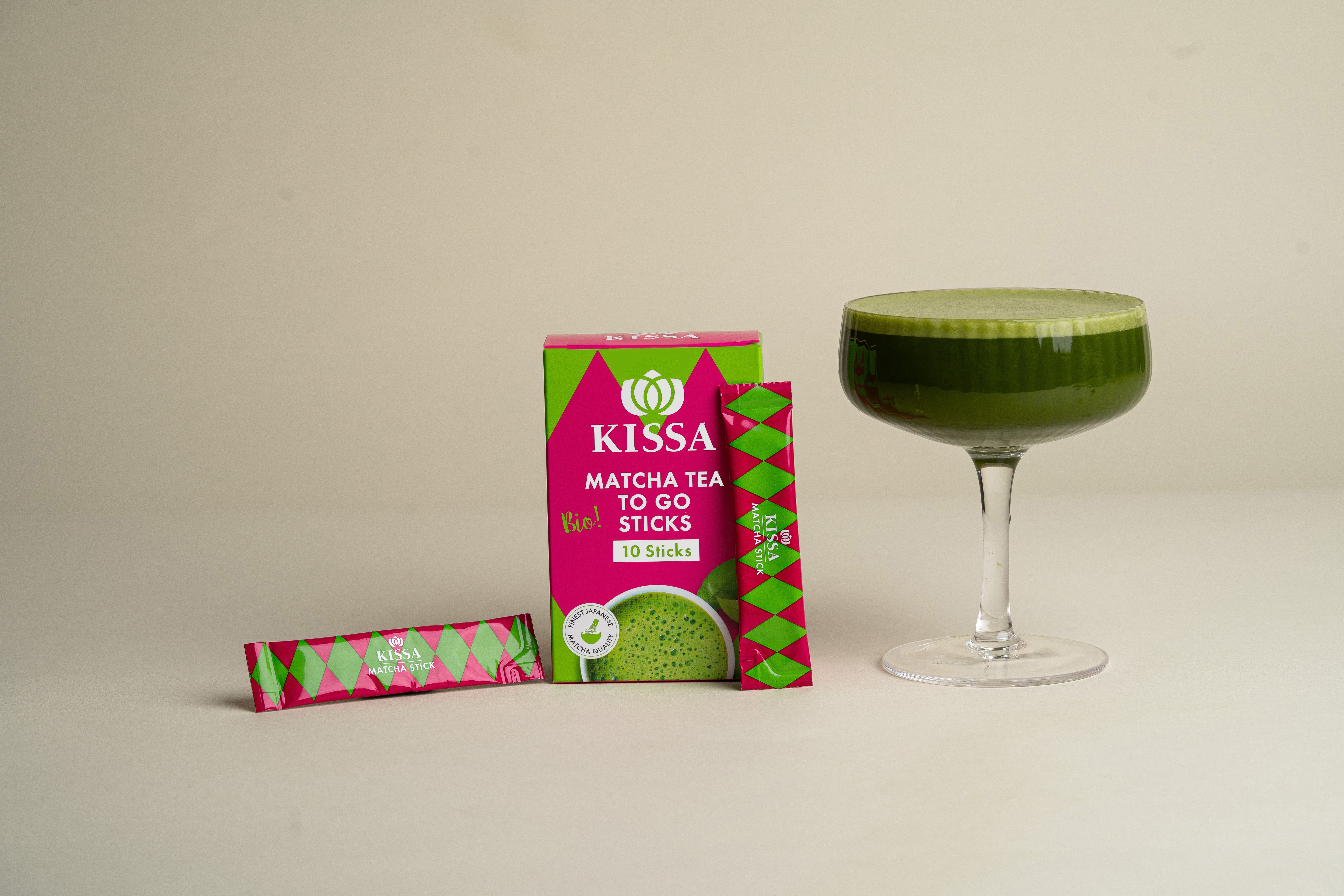 KISSA Matcha To Go Sticks in kompakter Box neben cremigem Matcha Latte auf hellem Hintergrund