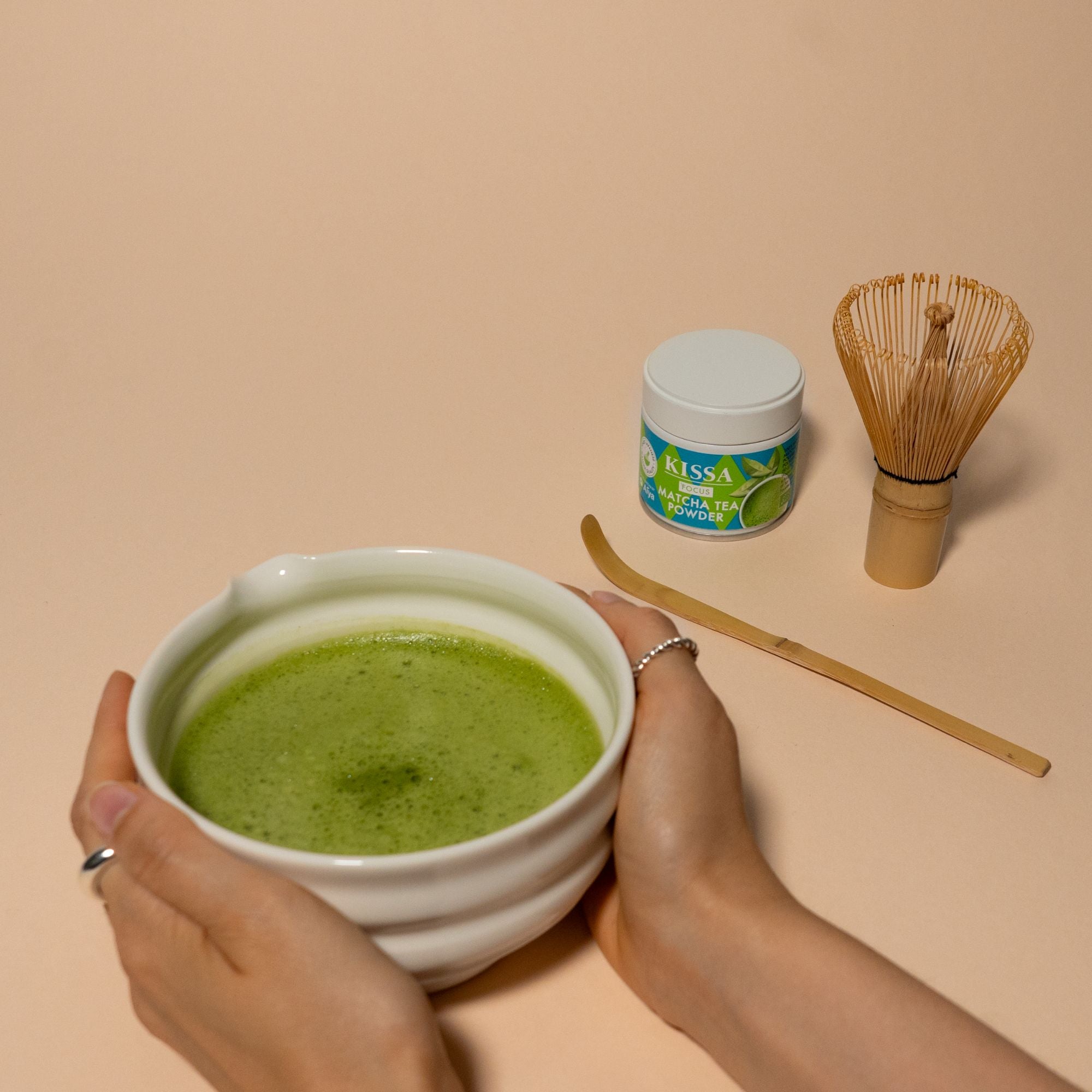 Hände halten Matcha-Schale mit reinem Matcha-Pulver, daneben ein Bambusbesen (Chasen), ein Bambuslöffel (Chashaku) und eine Dose KISSA Matcha Focus Tee Pulver.