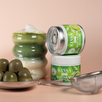 Two KISSA Matcha Basic tins next to a sieve, a Matcha Latte and Matcha Pralines