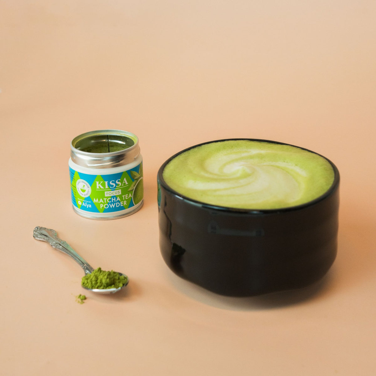 Black Miyabi matcha bowl filled with hot matcha and milk foam, placed next to a KISSA Matcha Focus tin, with a spoonful of matcha powder in front.