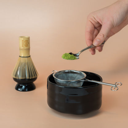 Black Miyabi matcha bowl with a matcha sieve, with a bamboo whisk resting on a black whisk holder behind it. Matcha powder is sprinkled from a spoon through the sieve.